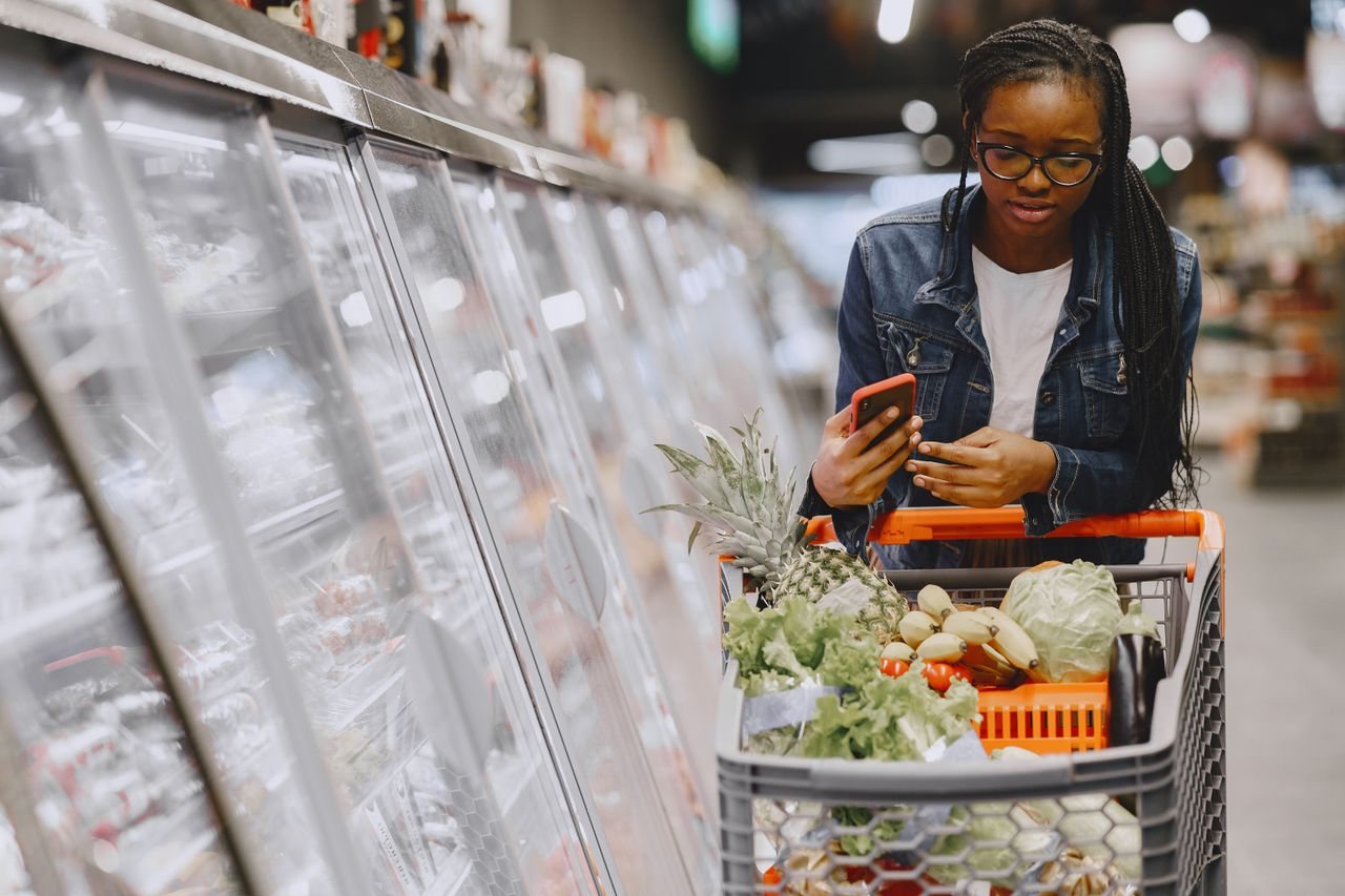 Free Woman in a grocery store using smartphone while pushing a shopping cart filled with groceries. Stock Photo