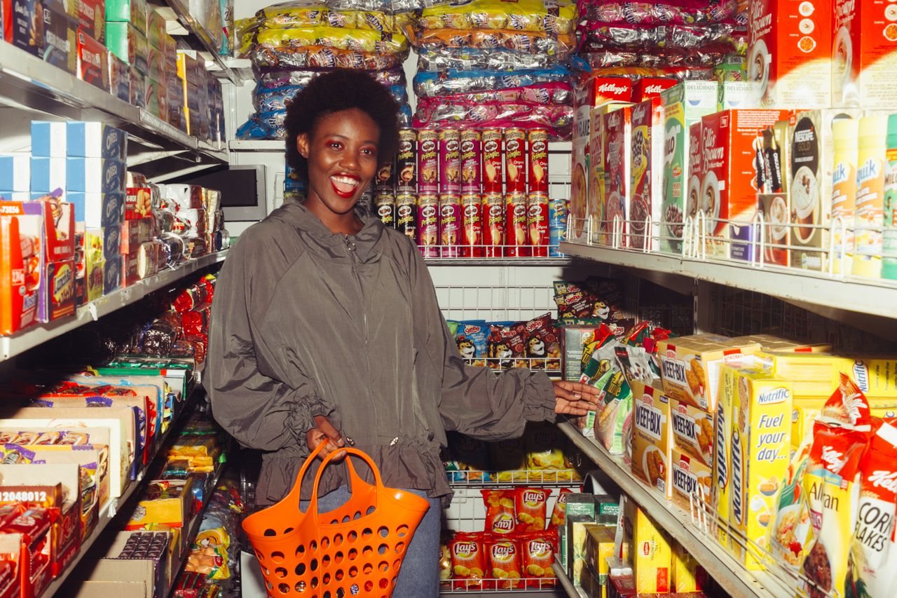 Free Happy African American lady in casual outfit with shopping basket in hand choosing food from products in small supermarket among colorful boxes on shelves and smiling with mouth opened Stock Photo